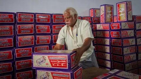 Getty Images A worker with white hair, wearing a cream coloured shirt, carries cartons of red match boxes to be loaded onto a cargo truck at a matchbox factory in Tamil Nadu, India. 
