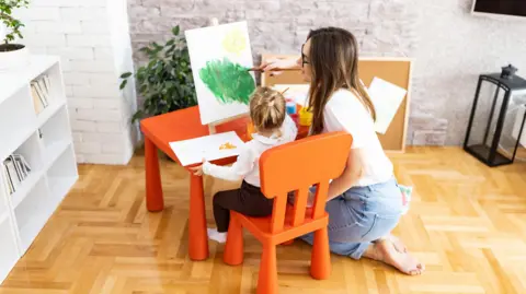 Getty Images A woman kneels on the floor next to a small child who is painting at a table 