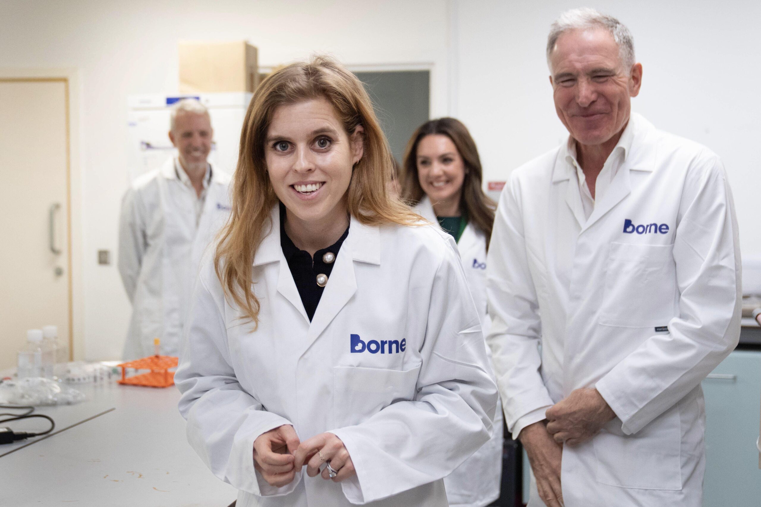 Princess Beatrice wearing a white lab coat and smiling with a group of people in lab coats behind her
