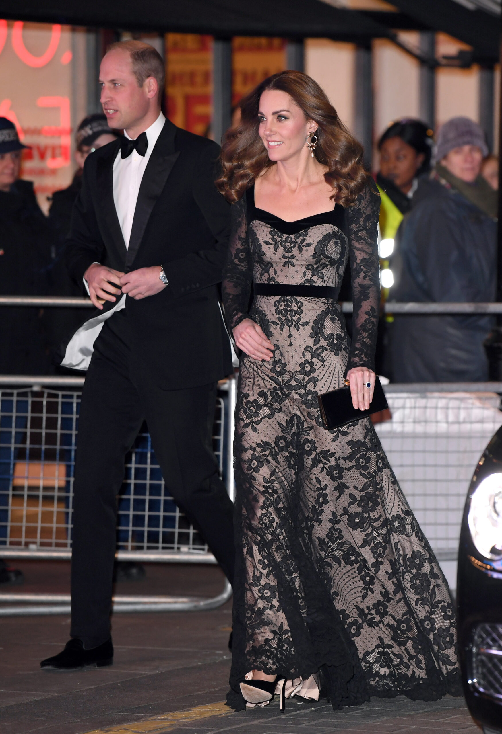 Prince William wearing a tux walking next to Kate Middleton in a black lace gown