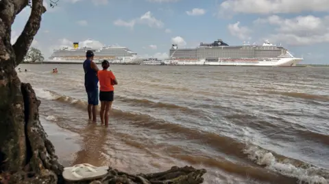 AFP via Getty Images Two people stand in the shallows of the river Guama and look out as two huge cruise ships are docked on the other side of the river against a blue cloudy sky. In the foreground a pair of white flip flops rest by a tree. Picture taken on 6 November.