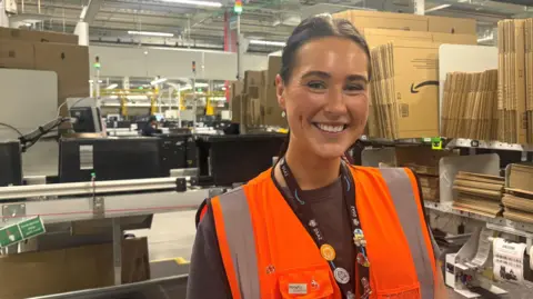 BBC A head and shoulders shot of Grace Rutter smiling at the camera. She is wearing a black t-shirt, an orange hi-vis vest and a black Amazon lanyard with badges on. She has long, dark brown hair which is tied back. She is standing in a warehouse. Cardboard boxes, a conveyer belt and machinery are behind her. 