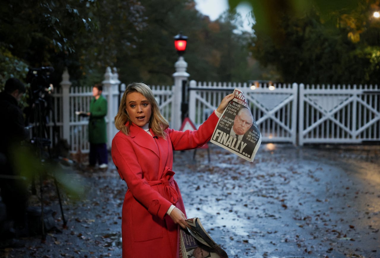 A person holds up a tabloid newspaper while standing in front of a gate.