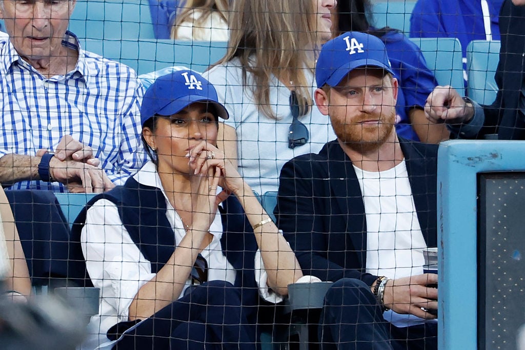 Two people wearing Los Angeles Dodgers caps sit in seats behind a protective netting.