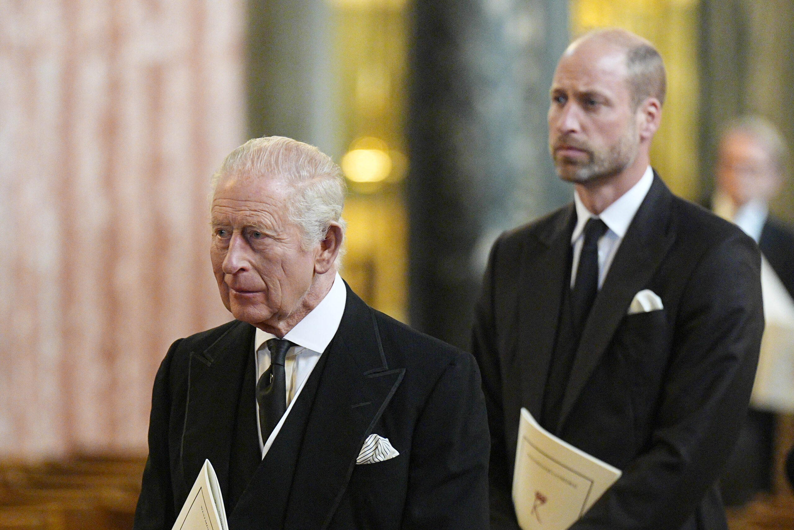 Prince William walking behind King Charles, both wearing black suits