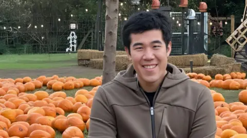 Yulin Wang Yulin Wang wearing a brown hoodie smiles while sitting in a park full of pumpkins.