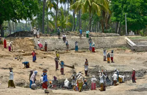 Hindustan Times via Getty Images) Women working for lake rejuvenation under MNREGA scheme at Bevanahalli village in Mandya, India. Pictured is a dry lake with women workers, with coconut plantations in the backdrop. 