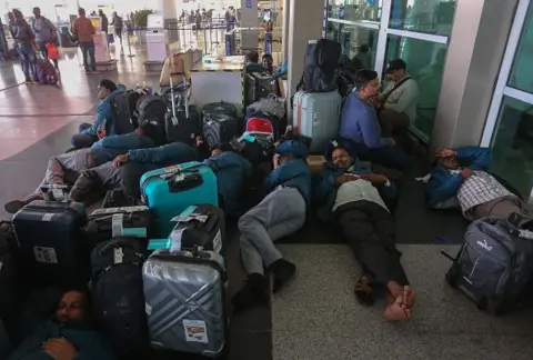 AFP via Getty Images Passengers rest outside of Terminal 1 of Indira Gandhi International Airport after mass cancellation of Indigo flights on December 05, 2025 in New Delhi, India. IndiGo, India's largest airline, cancelled over 200 flights across major hubs including Delhi, Mumbai, Bengaluru and Hyderabad in early December due to the implementation of new crew duty time regulations, leaving thousands of passengers stranded and frustrating travellers with minimal prior notification. 
