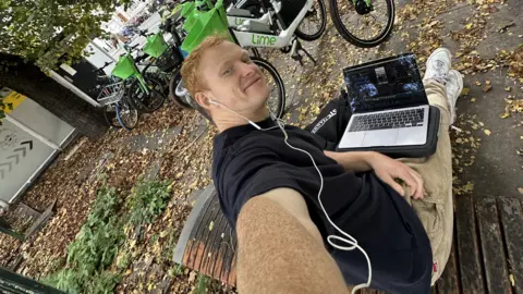 Sol Hyde Sol Hyde takes a selfie. He is sitting on a bench with his laptop. He is wearing a black T-shirt, khaki trousers and white trainers. He has white earphones in. There are autumn leaves on the ground and a number of Lime hire bikes behind him.
