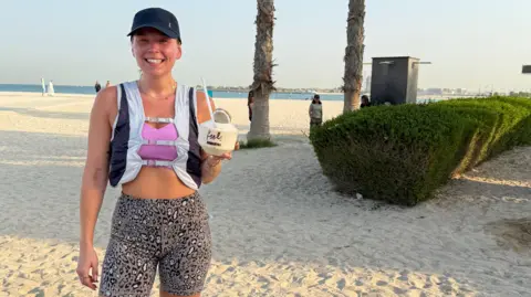 Isobel Perl Isobel, wearing running gear and a baseball cap, looks at the camera and smiles. She smiles at the camera and stands holding a coconut-shaped drink.