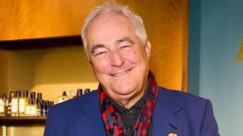 Lush Mark wearing a bright blue suit, patterned red-and-blue scarf. He is standing in a shop and behind him are wooden shelves displaying perfume bottles.