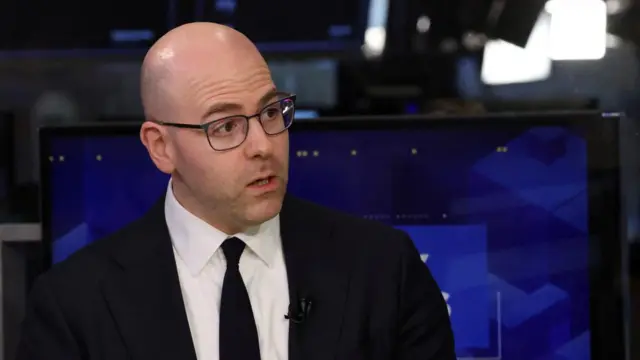 A bald man in glasses and a dark suit and tie looks to his left