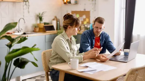 Getty Images A woman and her concerned-looking male partner sit at their kitchen table reading documents in front of a laptop. 