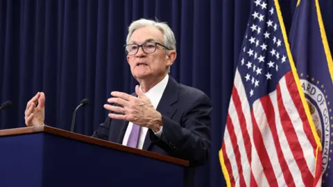 Reuters A man wearing a suit speaks in front of a podium. An American flag hangs in the background.