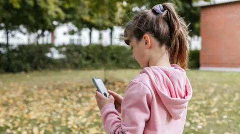 Getty Images A young girl with her hair in a ponytail wearing a pink hoodie looks at a phone