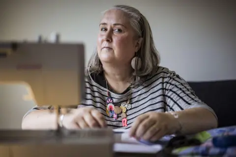 Chris Hopkins/BBC A woman wearing a striped t-shirt and colourful charm necklace sits behind a sewing machine, while fiddling with fabrics. She looks deep in thought.