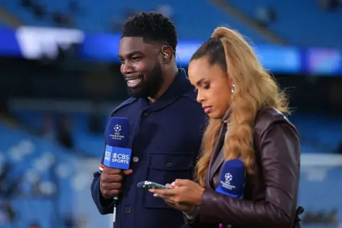 Danehouse/Getty Images Michah Richards stands next to a fellow female pundit holding a CBS Sports branded microphone in front of a football stand, mid-broadcast on the network.