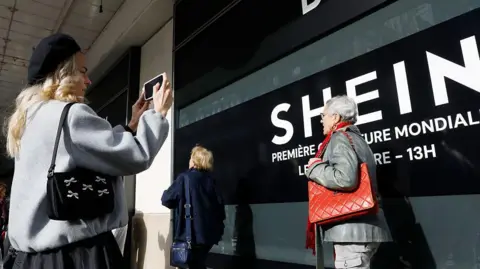 Getty Images A woman take a picture of a Shein banner at a Paris department store as an elderly woman carrying a red handbag walks by.