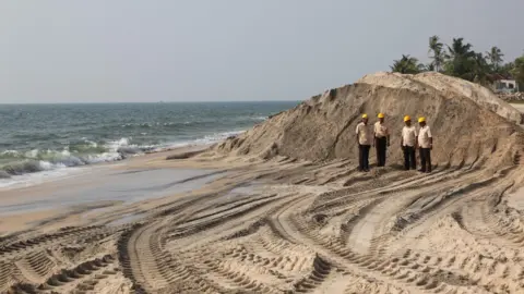 Corbis via Getty Images Four engineers wearing beige shirts and yellow helmets stand on the shores of a beach in Kerala where rare earth traces were found
