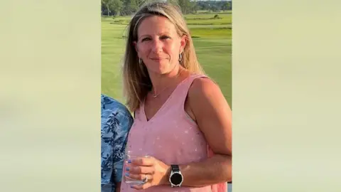 Kristy Hallowell A woman wearing a pink shirt holds a glass while posing in front of a green lawn.