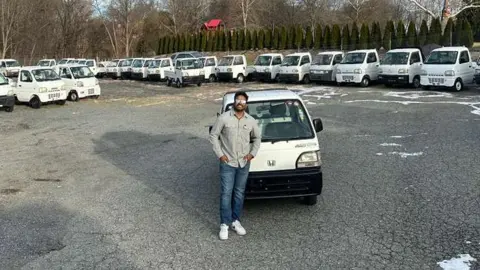 Tokyo Motors DC Mo Sulai posing with his hands in his pockets in front of a white Japanese mini truck in a car lot full of similar vehicles.  