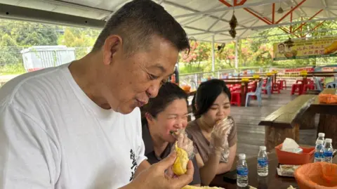 BBC/Koh Ewe Side view of a middle-aged man in a white T-shirt holding a slice of durian in one hand and a yellow glob of durian in the other hand. Behind him are two women sitting at a table eating durians with plastic gloves.