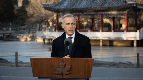 Getty Images Mark Carney, Canada's prime minister, speaking at a news conference in Beijing, China, on Friday, Jan. 16, 2026. He is wearing a black coat and tie, with a white button-up shirt, standing at a wooden podium with a maple leaf on its front side. Behind him is a backdrop of a water and a traditional Chinese structure in Beijing. 