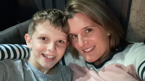 PA Media Jools (left) and Ellen (right) sit together in a chai, smiling for a selfie. Jools has short blonde hair and wears a grey top. Ellen has a blonde bob and wears a white, pink and navy striped jumper. 
