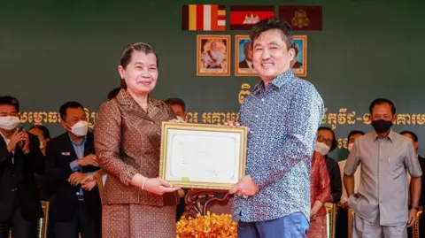 Kuong Li/Facebook A man and a woman both hold a framed certificate and smile at the camera, while a row of people wearing face masks stand behind them