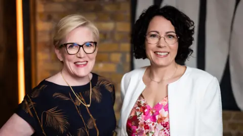 Piranha Photography With blonde hair and wearing a black blouse Monika Liikamaa  stands next to Denise Johansson who wears a white jacket and flowery pink blouse. 