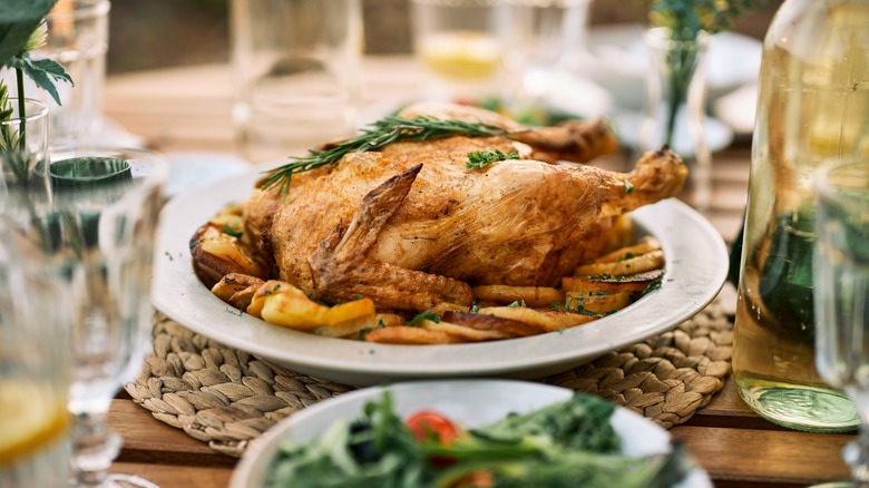 A beautifully set outdoor table with a golden roasted chicken surrounded by vegetables, fresh salad, and glasses, evoking a cozy meal